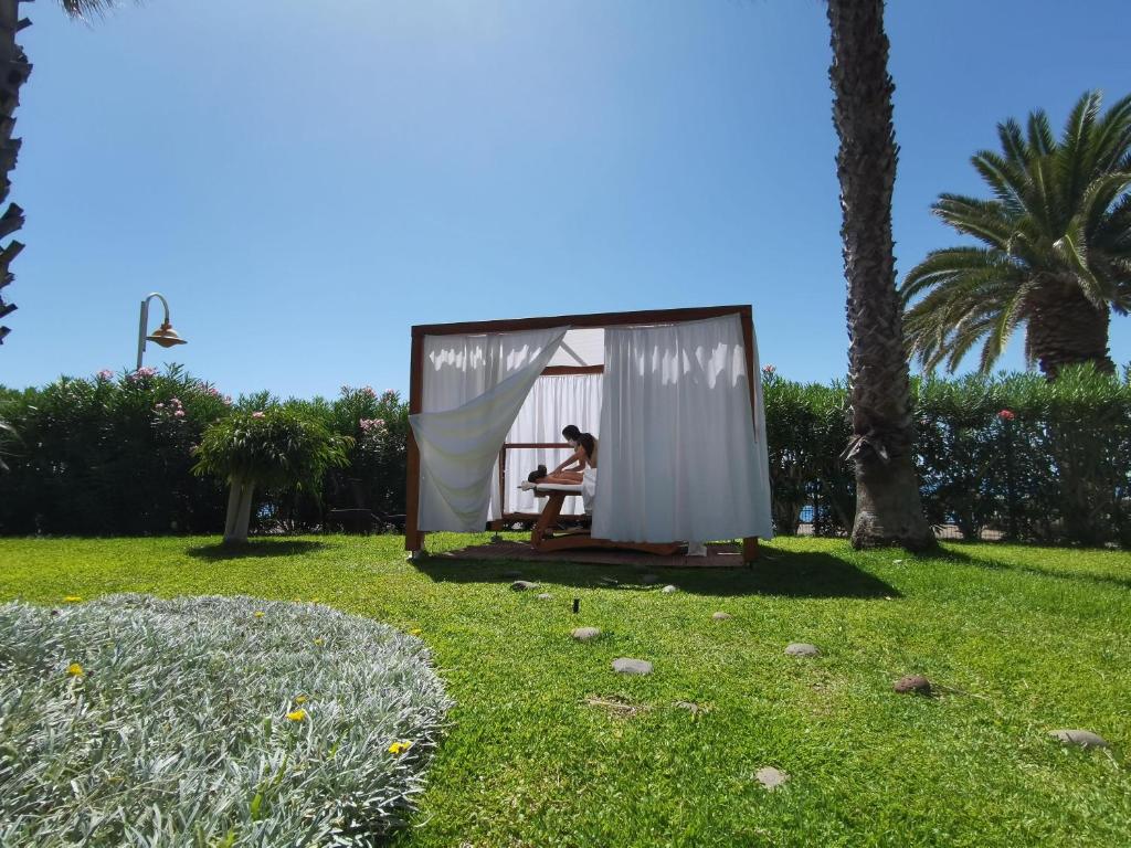 Atpūtas zona ar koka struktūru un baltiem aizkariem, kurā atrodas persona, kas relaksējas. Apkārt ir zaļš zāliens un palmas, radot mierīgu atmosfēru.