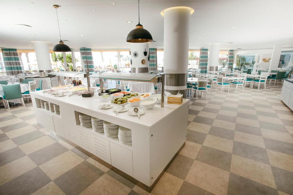 Gaismas un moderns restorāns ar ēdienu uzklāšanu, kurā redzams bufetes galds ar dažādiem dārzeņiem un augļiem, kā arī plaša ēdamzāle ar gaišām mēbelēm.