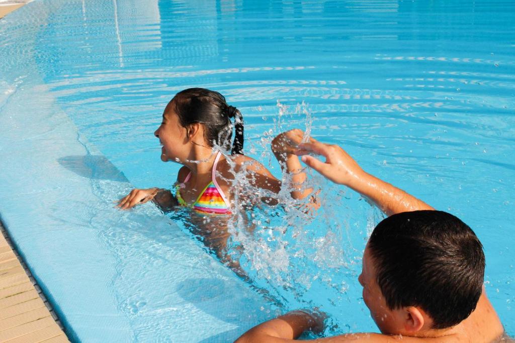 Divi bērni spēlējas baseinā, izšļakstot ūdeni un smejoties. Viens no bērniem ir tērpies krāsainā peldkostīmā, bet otrs ir tuvāk malai, radot jautru atmosfēru.