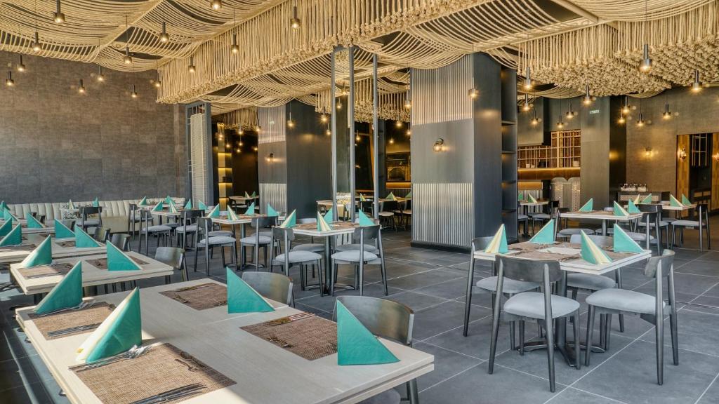 Mūsdienīgs restorāns ar eleganti iekārtotām galdiem, uz kuriem ir zilganzaļas salvetes. Gaismekļi un tekstils piešķir telpai siltu atmosfēru.