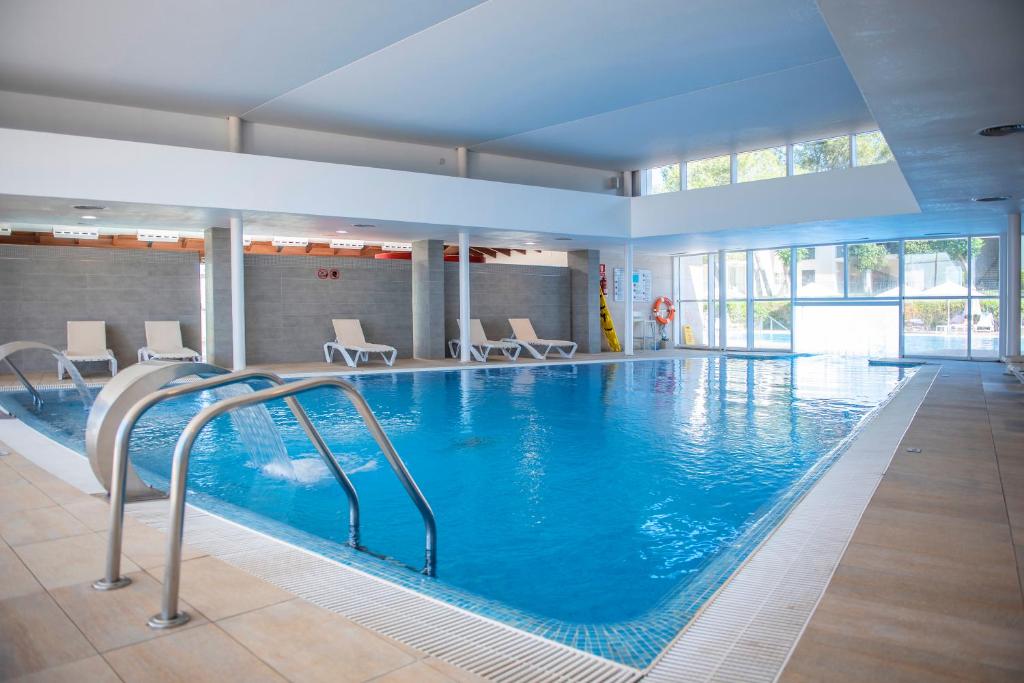 Iekštelpu peldbaseins ar skaidru ūdeni un sauļošanās krēsliem ap to, apgaismots ar dabiskiem gaismas avotiem.
