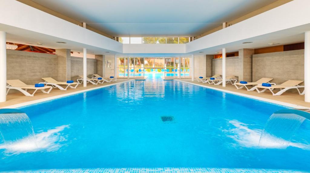 Mūsdienīgs iekštelpu peldbaseins ar skaidru zilu ūdeni un relaksējošām ūdens plūsmām, apkārtējo telpu piepilda saulaina gaisma un ērtas sauļošanās krēsli.