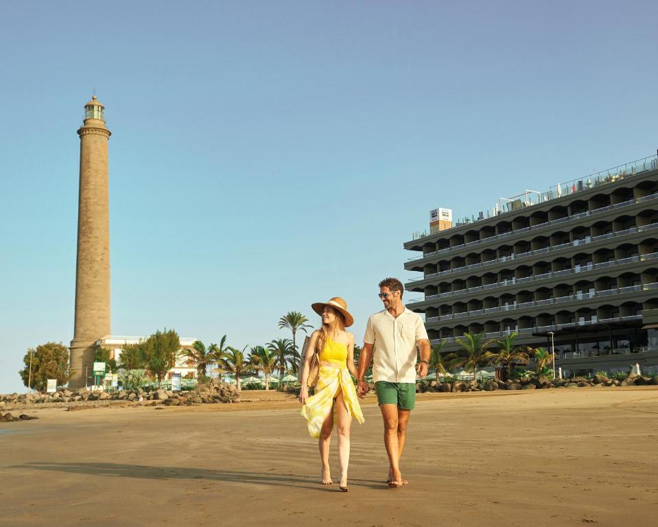 Pāris pastaigā gar pludmali, fonā redzams gaismas bāka un viesnīca. Šī aina iemieso romantisku atpūtu pie jūras.