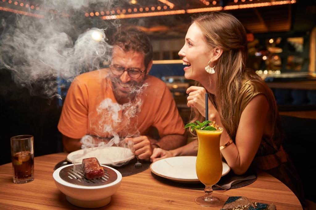 Divi cilvēki izbauda vakariņas modernā restorānā, kurā redzams tvaiks no gaļas ēdiena. Uz galda ir dzērienu glāzes un izsmalcināts kokteilis ar piparmētru.