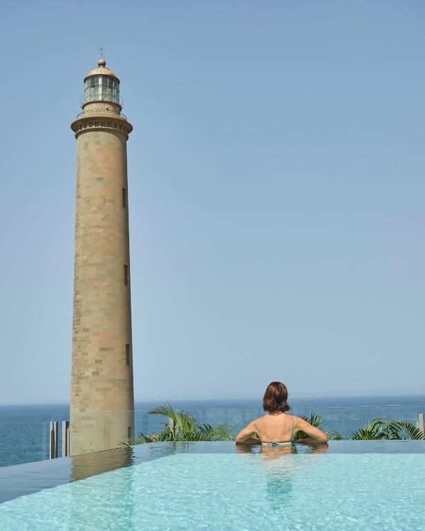 Skats no baseina uz jūras bākas, kur sieviete relaksējas pie bezgalības baseina. Skats uz mierīgu jūru un skaidru debesi.