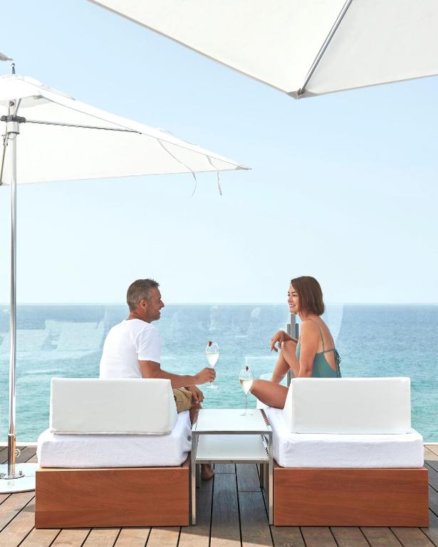 Pāris relaksējas uz terases ar skatu uz jūru, baudot kokteiļus. Apsildāmi sauļošanās krēsli un saulessargi rada ideālu atmosfēru atpūtai.