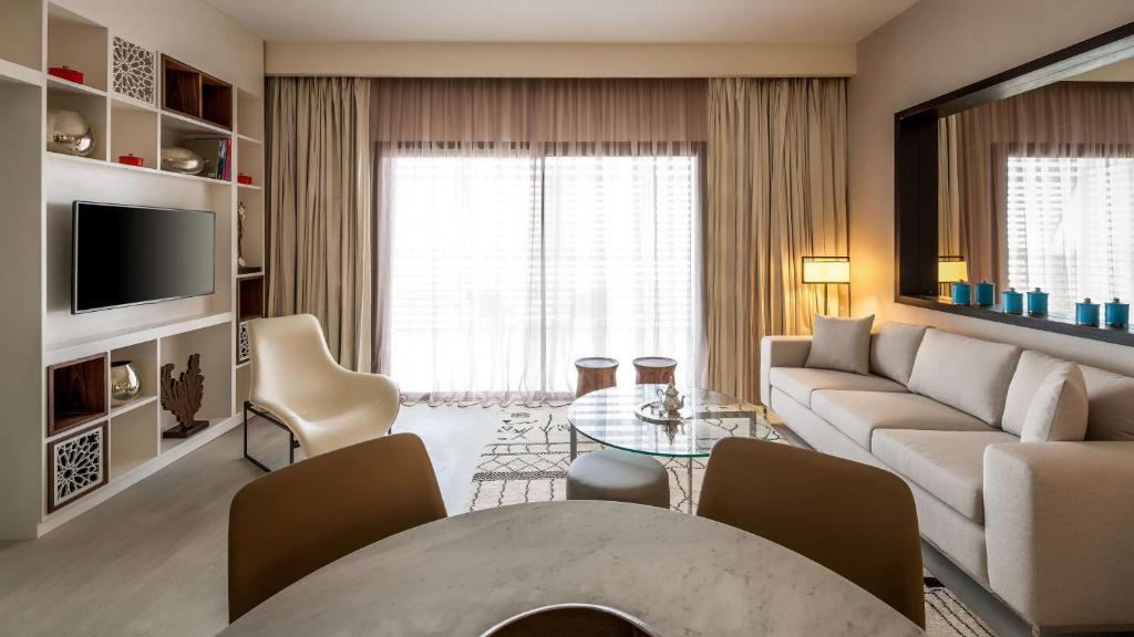 Mūsdienīgs viesistabas interjers ar gaišām krāsām, modernu mēbelēm un plašiem logiem, kas nodrošina dabisko apgaismojumu. Dzīvojamā zona ir aprīkota ar dīvānu, stikla galdu un televizoru.