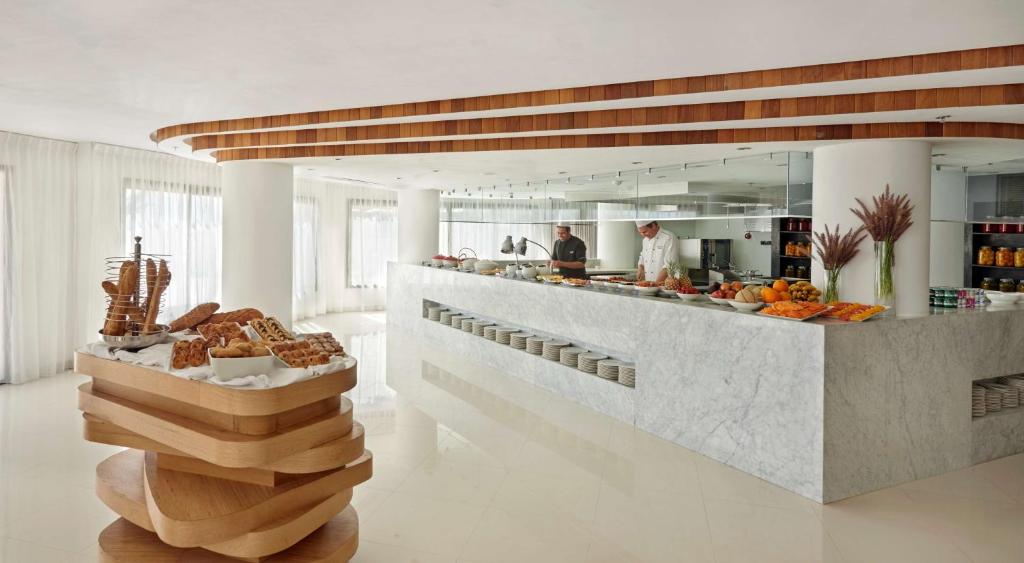 Mūsdienīga restorāna iekārtojums ar marmora letēm un koka elementiem, kur divi pavāri strādā virtuvē, bet priekšplānā ir dažādi maizes izstrādājumi un augļi.