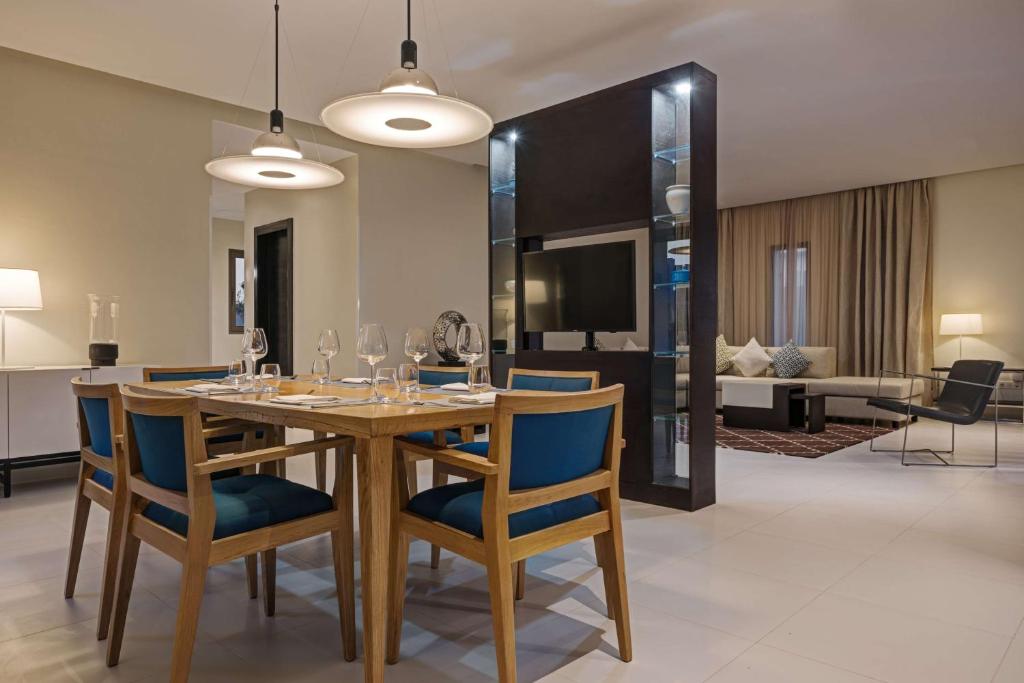 Mūsdienīga viesistaba ar ēdamgaldu, koka krēsliem un stilīgu apgaismojumu. Interjerā ir redzams televizors un dīvāns, radot ērtu un viesmīlīgu atmosfēru.