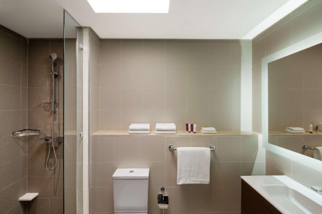 Mūsdienīgs vannas istabas interjers ar stikla dušu, baltiem dvieļiem un elegantu apgaismojumu. Vannas istabā ir arī moderna izlietne un tualete.