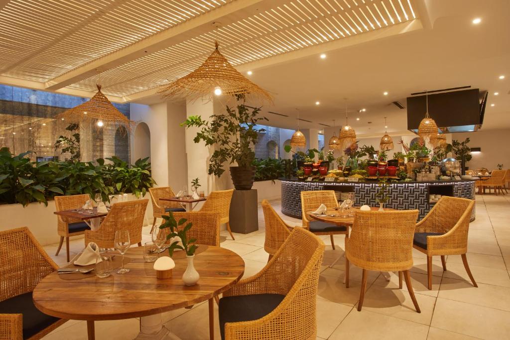 Mūsdienīgs restorāns ar zaļām augu dekorācijām, rattan krēsliem un elegantu apgaismojumu, radot patīkamu un relaksējošu atmosfēru.