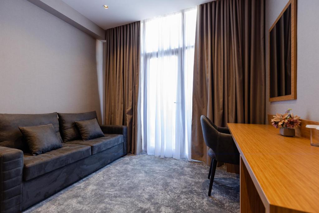 Mūsdienīgs viesnīcas numurs ar pelēku dīvānu, koka galdu un gaišu logu ar aizkariem. Telpa ir eleganta un ērta, ideāla relaksācijai.