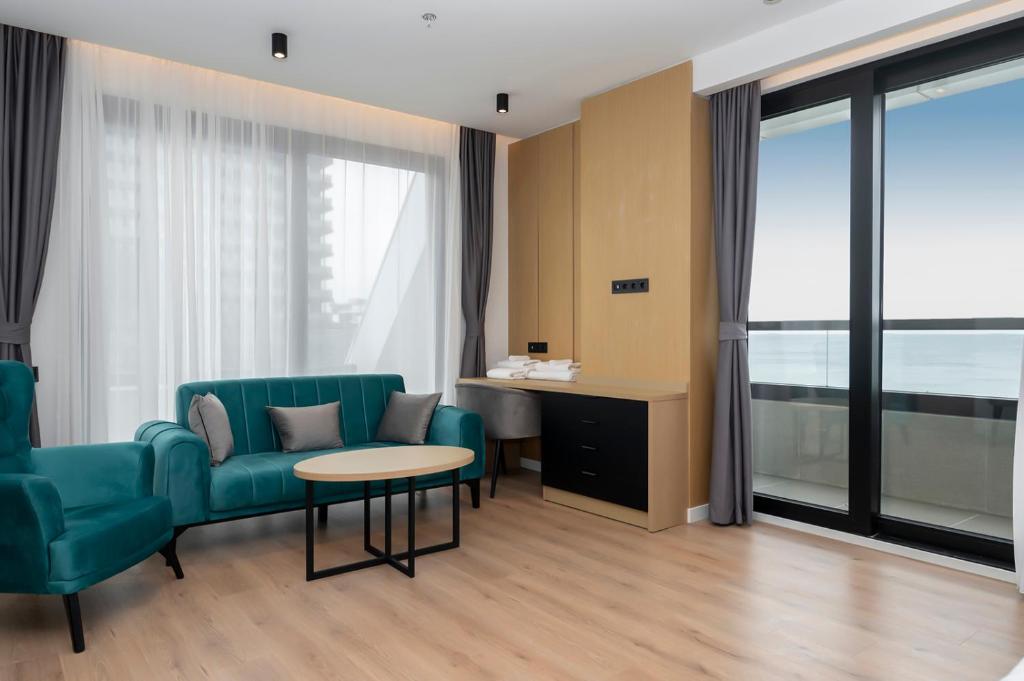 Mūsdienīgs viesnīcas numurs ar teal krāsas mēbelēm, koka grīdu un panorāmas logiem, kas piedāvā skatu uz jūru. Interjers ir gaišs un elegants, radot relaksējošu atmosfēru.