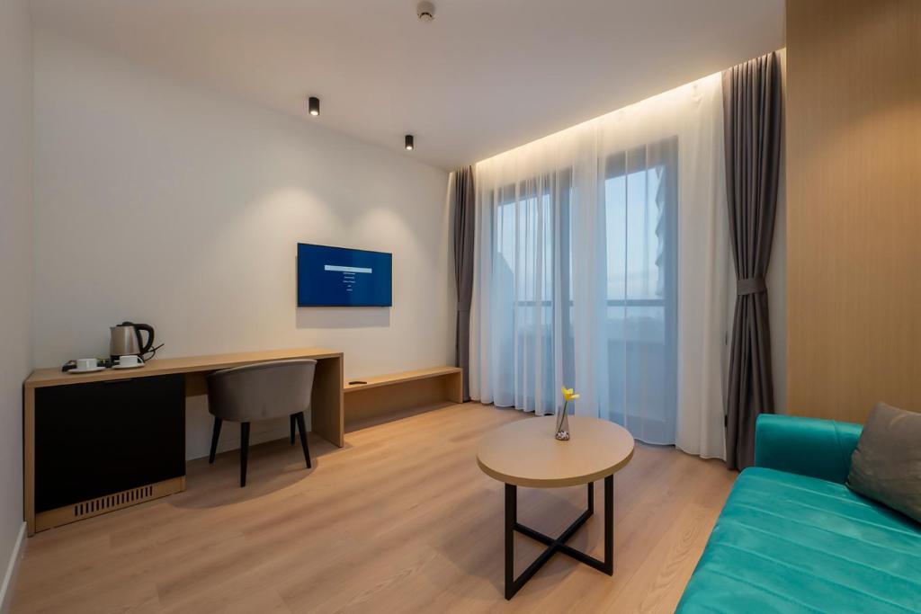 Mūsdienīgs viesistabas interjers ar dīvānu, galdu un televizoru, kas atrodas pie lieliem logiem ar aizkariem. Uz galda ir zieds un pie sienas ir rakstāmgalds ar krēslu.