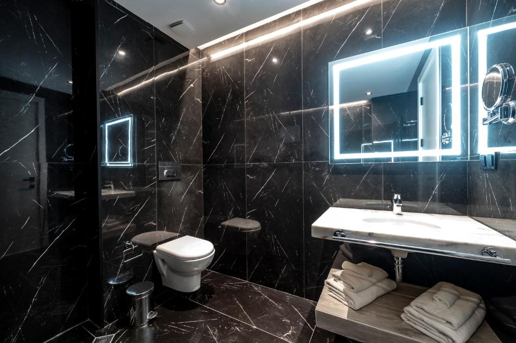 Mūsdienīgs vannas istabas interjers ar melnu marmora apdari, apgaismotu spoguli un elegantu izlietni. Vannas istabā ir arī tīra tualete un uzglabāšanas vieta dvieļiem.