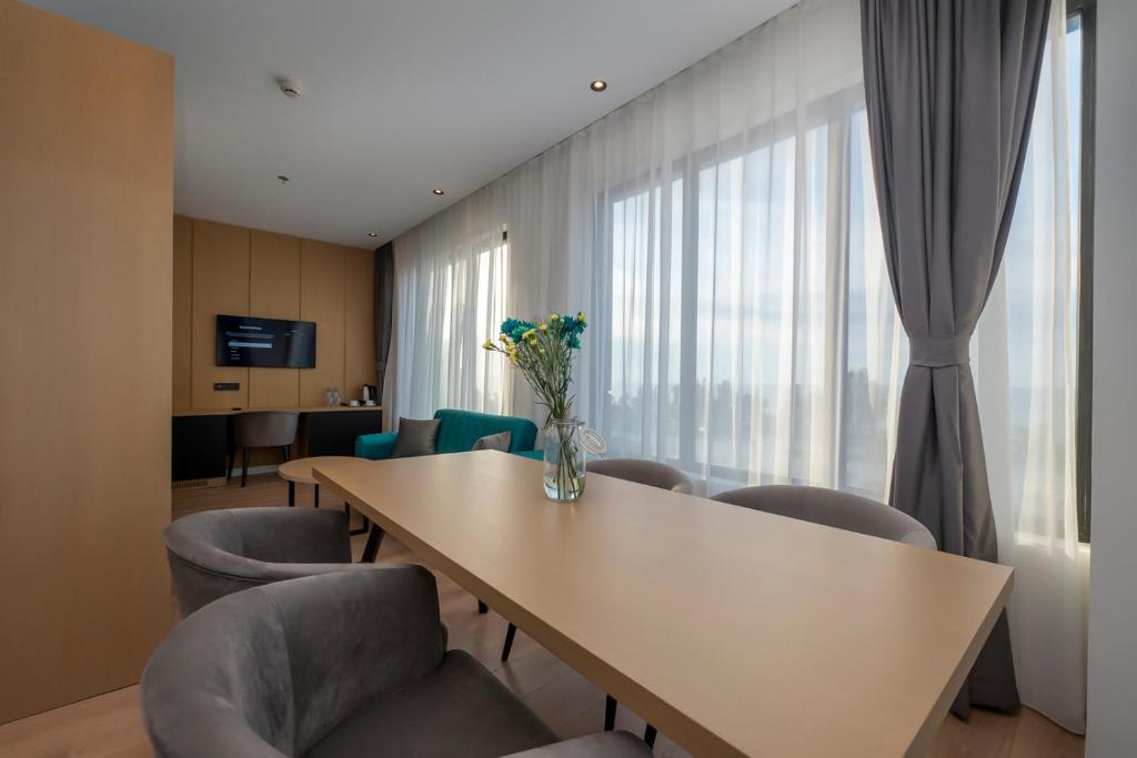 Mūsdienīgs viesnīcas numurs ar plašu logu, koka mēbelēm un elegantu apgaismojumu, kurā redzams galds ar ziediem un ērti krēsli.