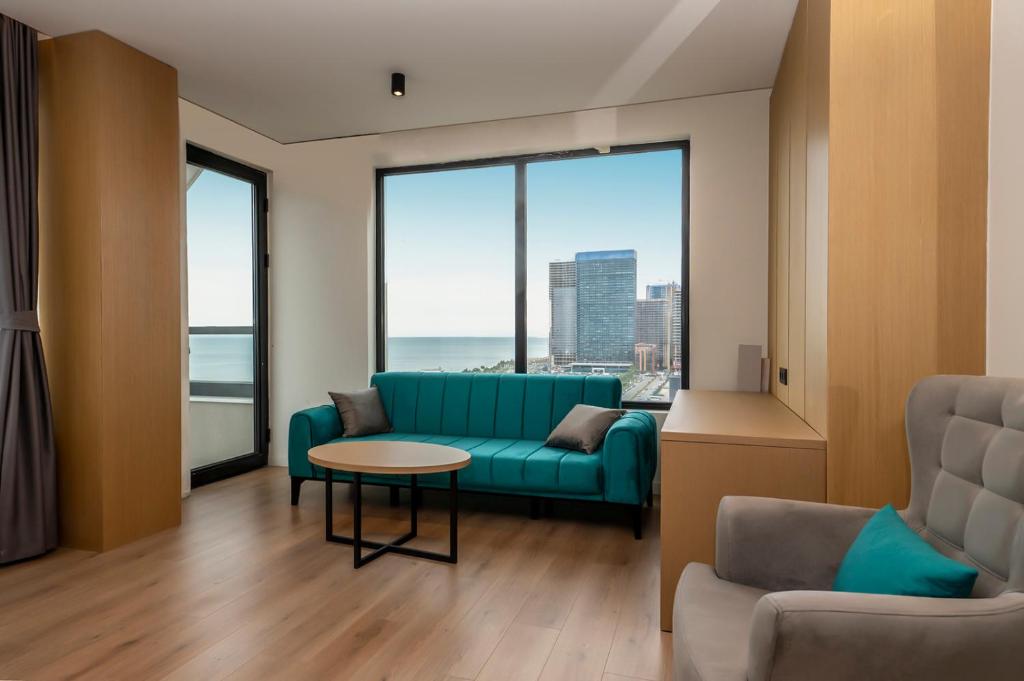 Mūsdienīgs dzīvojamās telpas interjers ar skatu uz jūru, kurā ir teal krāsas dīvāns, apaļs galds un gaišas koka grīdas. Logi sniedz plašu skatu uz pilsētu un ūdeni.