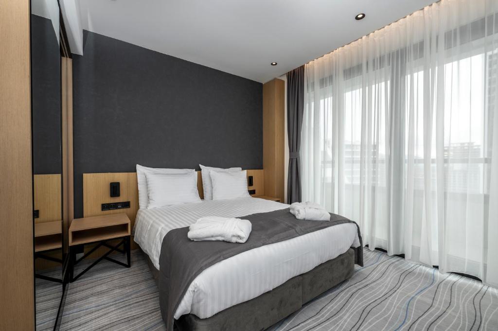 Mūsdienīga viesnīcas istaba ar divguļamo gultu, mīkstiem spilveniem un lieliem logiem, caur kuriem ieplūst dabiska gaisma. Interjers ir elegants ar koka un tumšu sienu akcentiem.