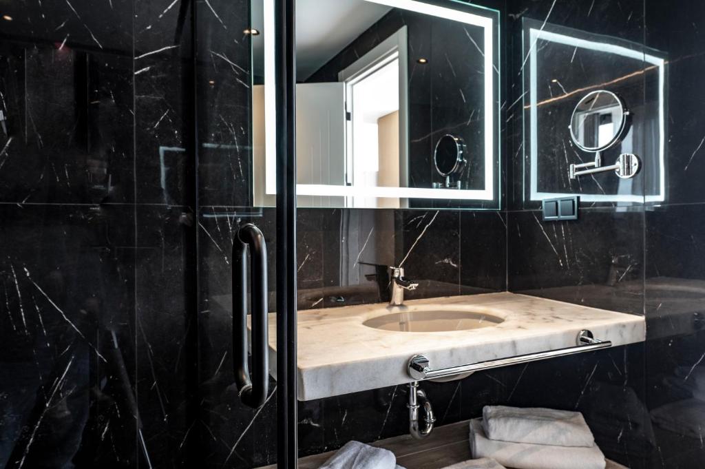 Mūsdienīgs vannas istabas interjers ar melnu marmora sienām, apgaismotu spoguli un elegantu izlietni. Svaigi salocītas dvieļi un mūsdienīgas santehnikas detaļas papildina telpas dizainu.