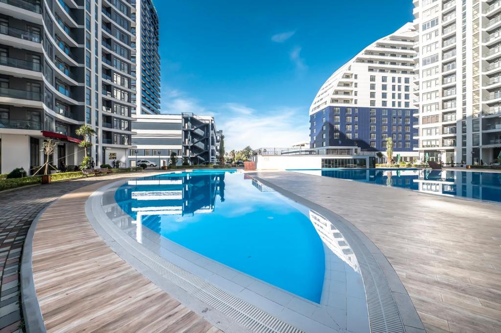 Mūsdienīga dzīvojamā kompleksa baseins ar skaidru, zilu ūdeni un plašu terasi, kas ieskauts ar augstceltnēm un zilo debesu fonu.
