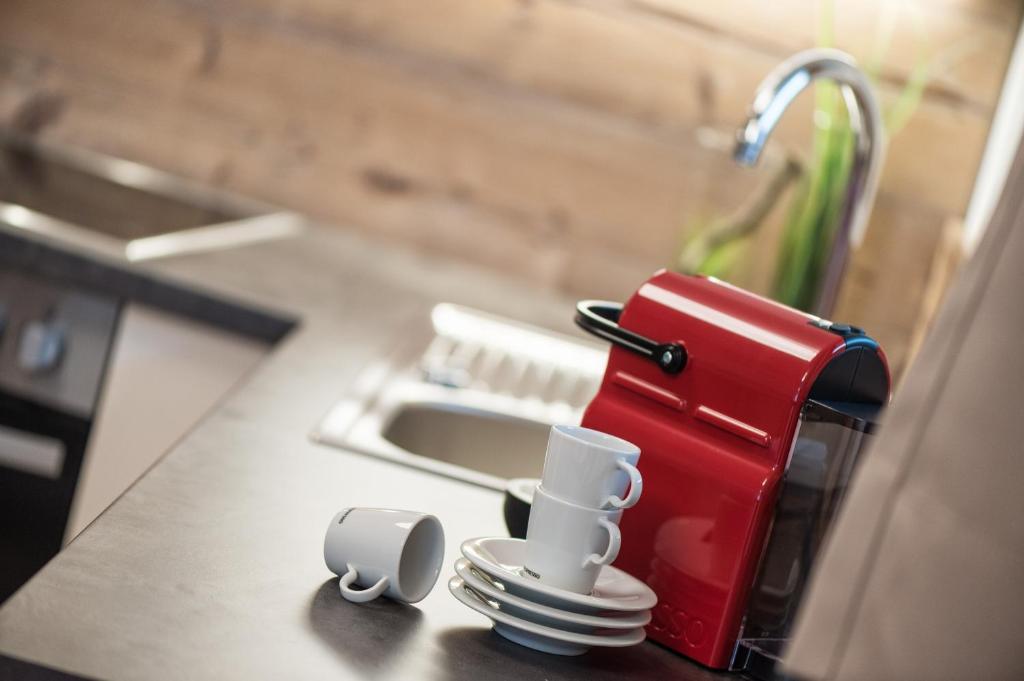 Sarkans kafijas automāts uz virtuves galda ar baltām krūzēm un šķīvjiem. Interjers rada mājīgu atmosfēru.