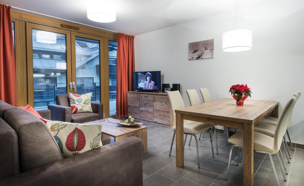 Mūsdienīga viesistaba ar koka mēbelēm, ērtu dīvānu un ēdamgaldu, kas rotāts ar ziediem. Telpā ir lieli logi un televizors, radot patīkamu atmosfēru.