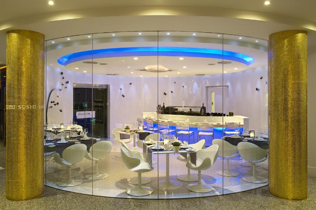 Moderns sushi restorāns ar stiklotu interjeru, elegantiem galdiem un krēsliem, kas apgaismots ar zilu gaismu. Svaiga un stilīga atmosfēra.