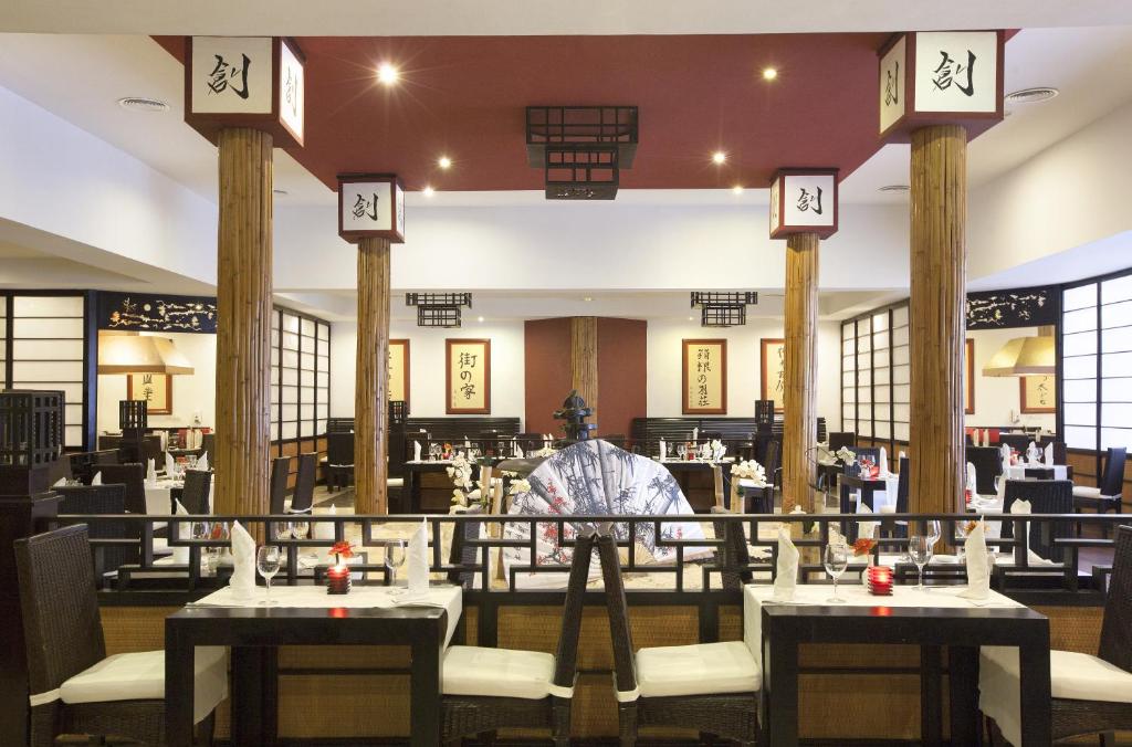 Mūsdienīga japāņu restorāna iekšskats ar elegantiem galdiem, tradicionāliem elementiem un mājīgu atmosfēru. Interjerā ir redzami koka stabi un dekoratīvi akcenti, kas rada autentisku noskaņu.