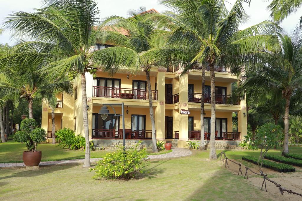 Tropu viesnīca ar palmām un dārzu, kas attēlo trīs stāvu ēku ar plašām terasēm un balkoniem. Viesnīcas nosaukums 