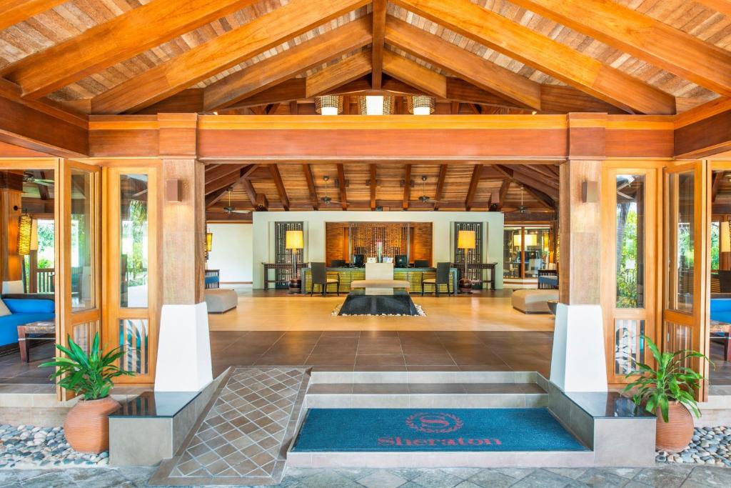 Mūsdienīgs viesnīcas vestibils ar koka konstrukciju un plašu telpu, kas piedāvā ērtas sēdvietas un zaļus augus. Viesnīcas logotips ir redzams pie ieejas.