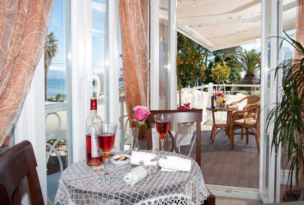 Eleganti iekārtota ēdamistaba ar skatu uz jūru, kur uz galda ir pudele vīna, glāzes un rozes zieds. Aiz loga redzamas palmas un saulaina terase.
