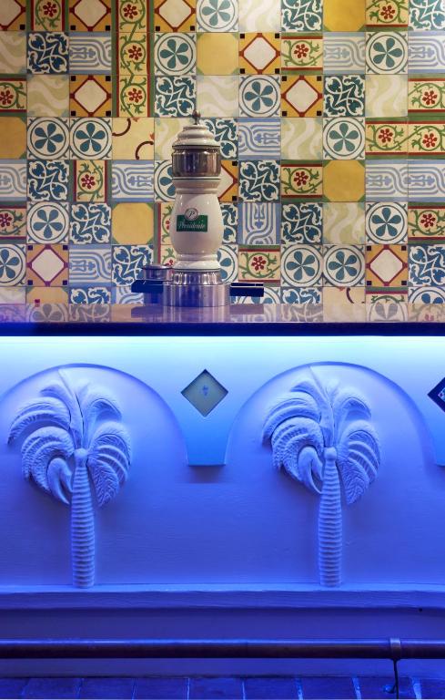 Krāsaina flīžu siena ar ziedu un ģeometriskiem rakstiem, centrā ir stilīgs dzērienu aparāts ar baltu un sudraba krāsu. Apakšā redzama eleganta apdare ar palmu motīviem un zilu apgaismojumu.