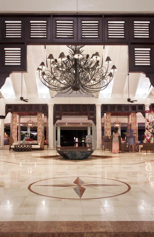 Elegants viesnīcas vestibils ar izsmalcinātu lustru un marmora grīdu, centrā ar dekoratīvu baseinu. Apgaismojums rada siltu un viesmīlīgu atmosfēru.