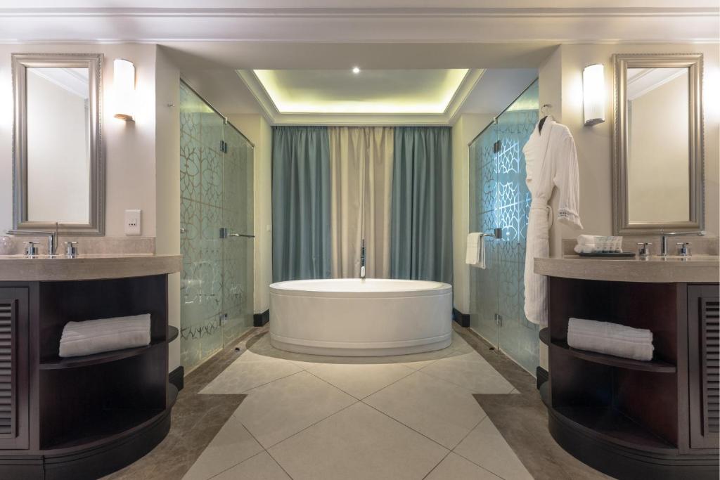 Mūsdienīgs vannas istabas interjers ar brīvu stūra vannu, stikla dušu un elegantiem spoguļiem, radot relaksējošu atmosfēru.