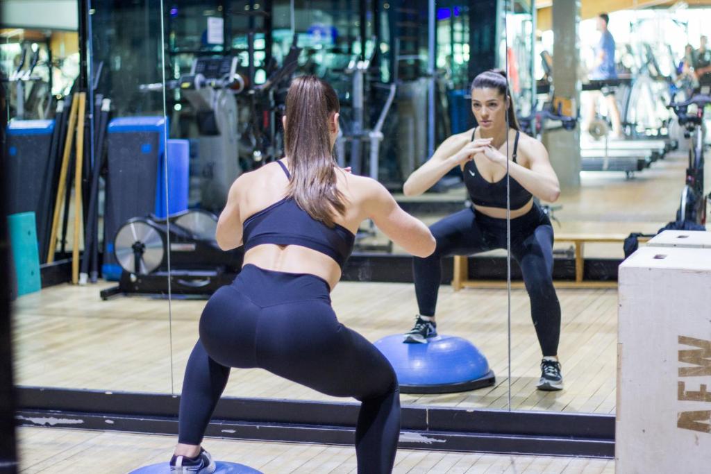 Sieviete veic squatu treniņu sporta zālē, skatoties spogulī. Fons ir aprīkots ar trenažieriem un sporta ekipējumu.