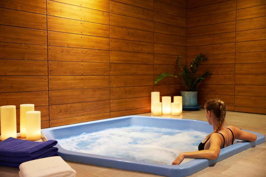 Mūsdienīgs spa ar zilu burbuļvannu un siltu apgaismojumu, kur blakus atrodas sveces un augi, radot relaksējošu atmosfēru.