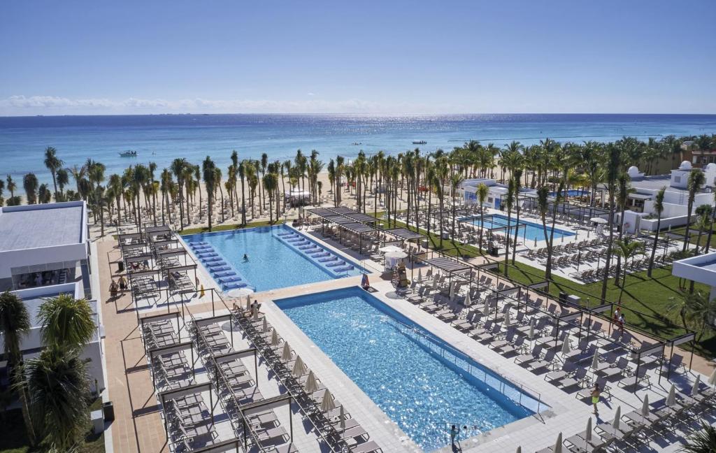 Viesnīcas komplekss pie pludmales ar diviem baseiniem un palmām, kas nodrošina atpūtu un relaksāciju. Skats uz jūru ar skaidru zilu debesi.