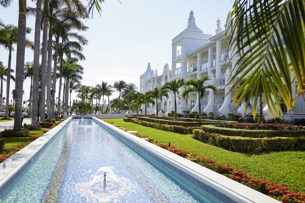 Eleganta viesnīca ar baltiem fasādēm, kas ieskauta palmām un krāšņiem dārziem, kur centrā plūst mierīgs ūdens kanāls.