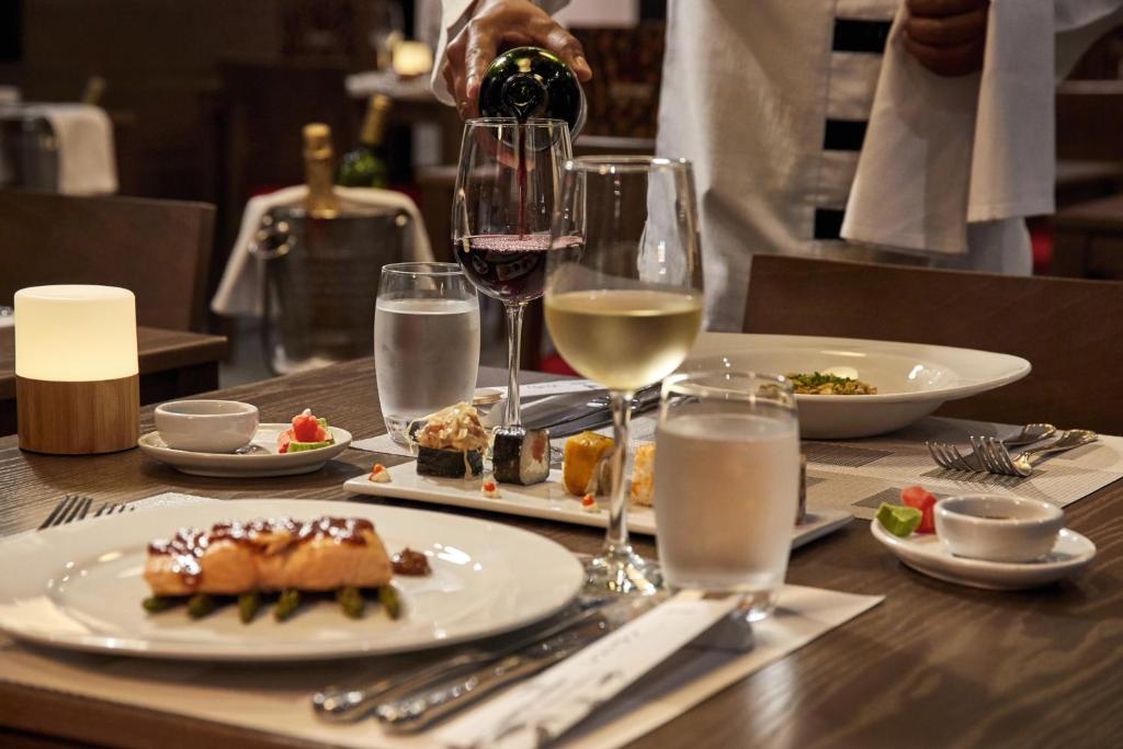 Elegants restorāna galds ar dažādiem ēdieniem, vīna glāzēm un ūdens traukiem, kam kalpo viesmīlis. Gaisma un atmosfēra rada patīkamu vakariņu noskaņu.