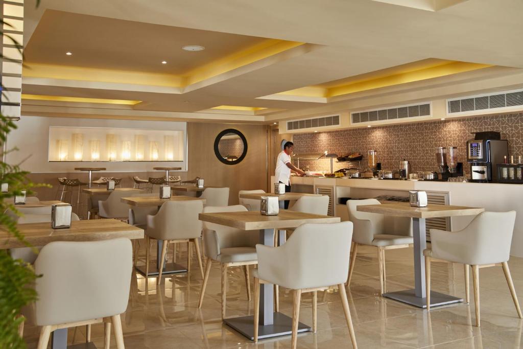 Mūsdienīgs restorāns ar gaišu interjeru un koka galdiem, kurā redzams apkalpojošais personāls, kas pasniedz ēdienus bufetē. Gaisma rada siltu atmosfēru, padarot to par patīkamu vietu ēšanai.