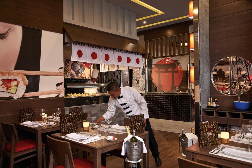 Svaigā un stilīgā japāņu restorāna interjers ar sushi gatavošanas zonu un viesmīli, kas sagatavo galdu. Restorānā redzami tradicionāli japāņu elementi un moderns dizains.