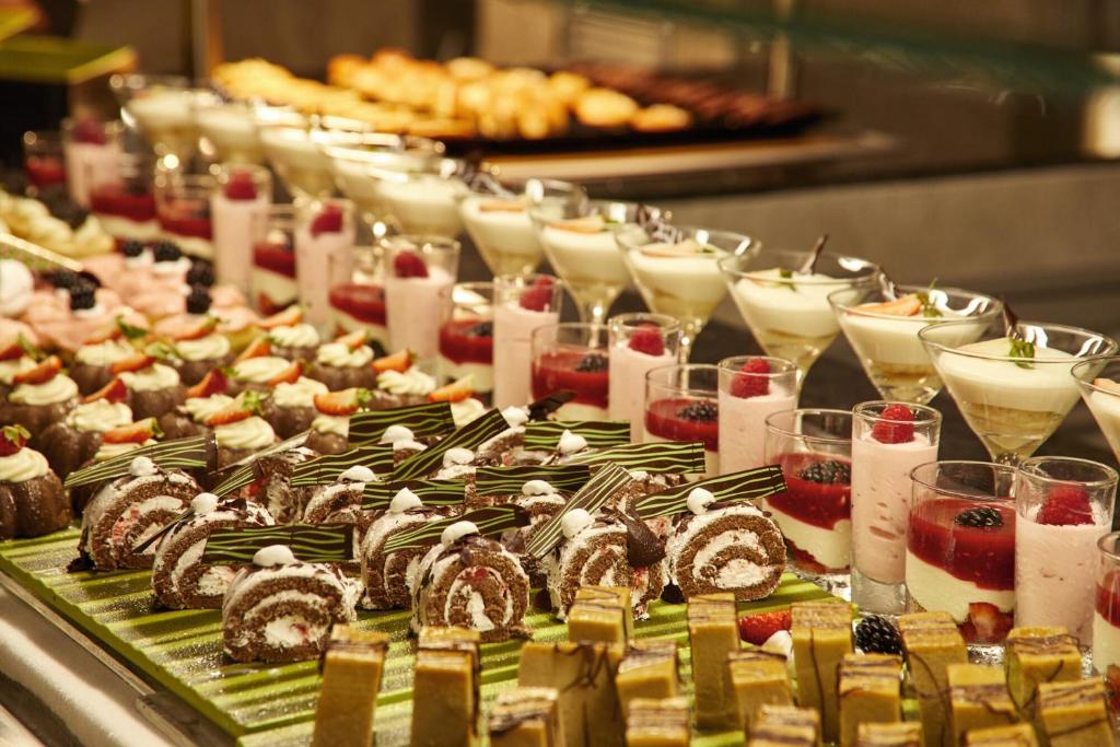 Izsmalcināts desertu bufete ar dažādiem saldumiem, ieskaitot šokolādes kūkas, krēmveida pudiņus un augļu desertus. Saldējumi un kūkas ir grezni sakārtoti, radot vilinošu un krāsainu skatu.