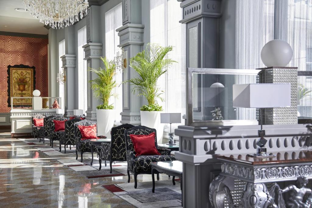 Elegants viesnīcas interjers ar grezniem krēsliem, spilveniem un sulīgiem zaļumiem, kas rada viesmīlīgu atmosfēru. Lielas logu ailes ļauj ienākt dabiskajai gaismai, uzsverot telpas šarmu.