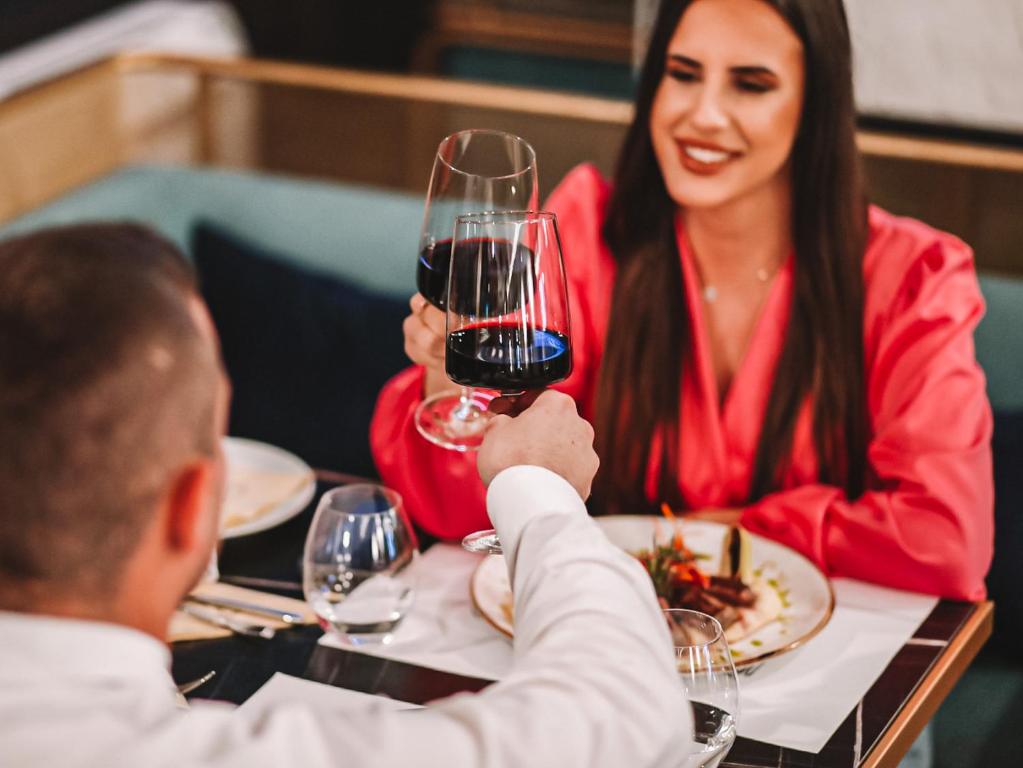 Romantiska vakariņas ar sarkanvīnu, kur pāris satiek un svin ar glāzēm. Fons ir elegants restorāns ar izsmalcinātu galda klājumu.