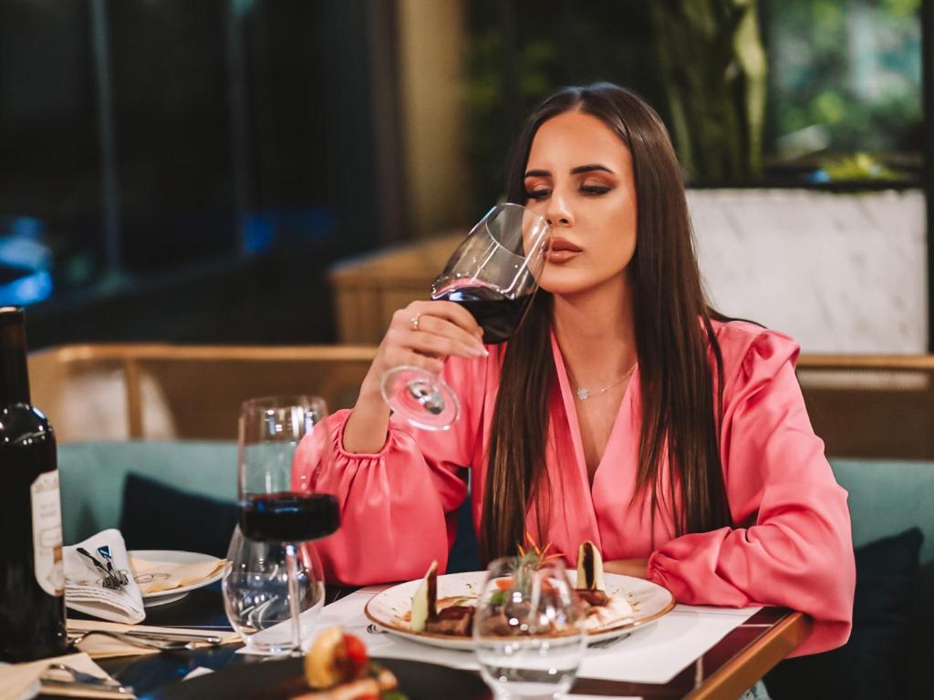 Sieviete rozā blūzē, baudot sarkanvīnu eleganti iekārtotā restorānā, ar izsmalcinātu ēdienu priekšā. Galda dekors un noskaņa rada intīmu un greznu atmosfēru.