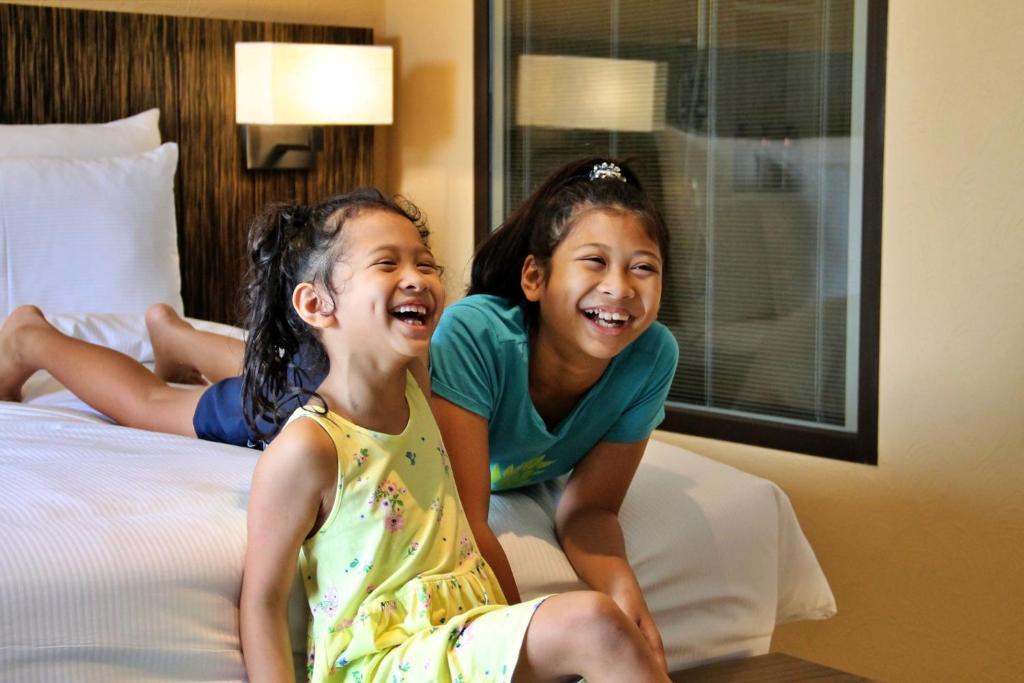 Divas laimīgas meitenes smejas viesnīcas numurā, sēžot uz gultas. Viņas izskatās priecīgas un jautras, baudot kopā pavadīto laiku.