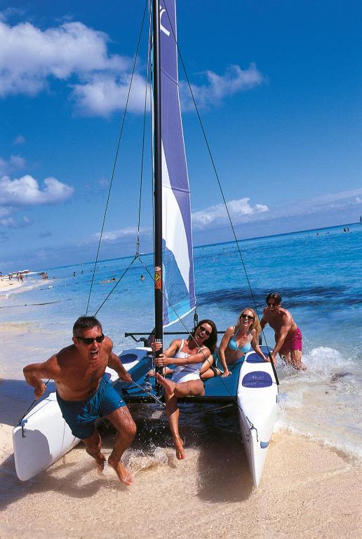 Četri cilvēki izbauda jūras burāšanu, stāvot uz katamarāna pie pludmales. Skaista saulaina diena ar zilo ūdeni un mākoņiem debesīs.