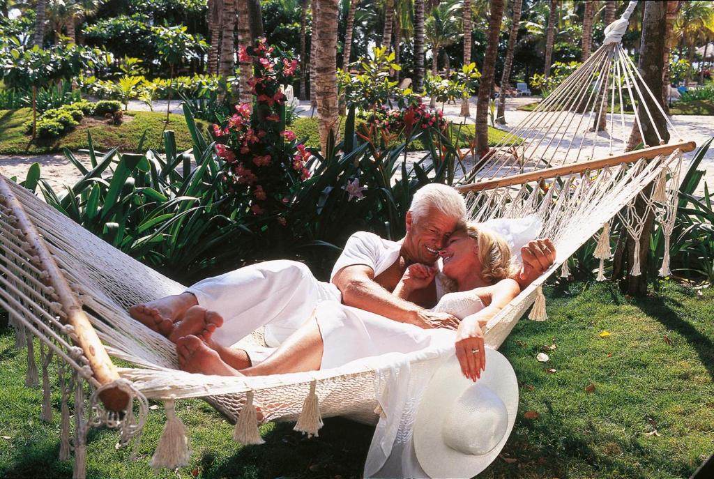 Skaists pāris atpūšas šūpuļtīklā tropu dārzā, izbaudot romantisku brīdi. Viņi ir ģērbušies baltās drēbēs, un apkārt redzams krāšņs ziedu un zaļumu fons.