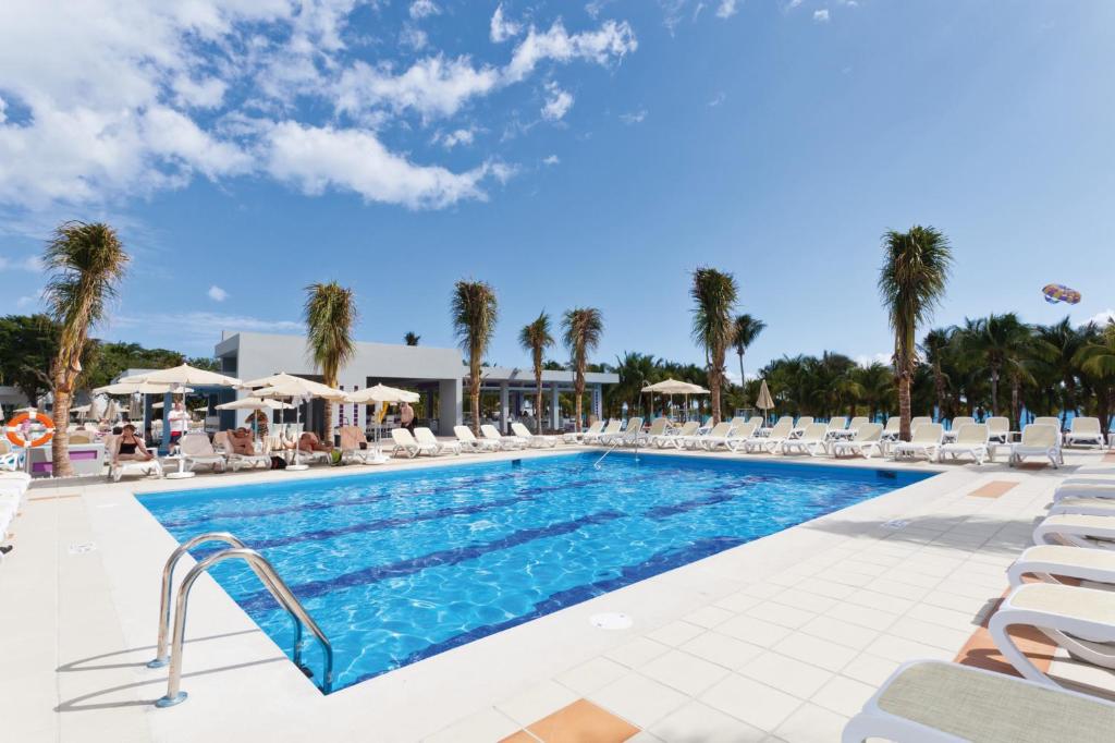 Baseins ar palmām un sauļošanās krēsliem, ideāls atpūtas vieta saulainā dienā. Apkārt ir tropiska vide un skaidra zila debesis.