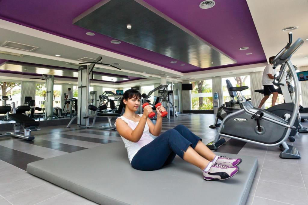 Sporta zāle ar modernām iekārtām, kur sieviete veic treniņu, turēdama hanteles rokās. Aktīva vide veicina veselīgu dzīvesveidu un fizisko sagatavotību.